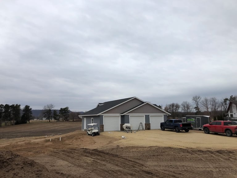 New residential home with two-car garage under construction in rural area with vehicles and bare ground
