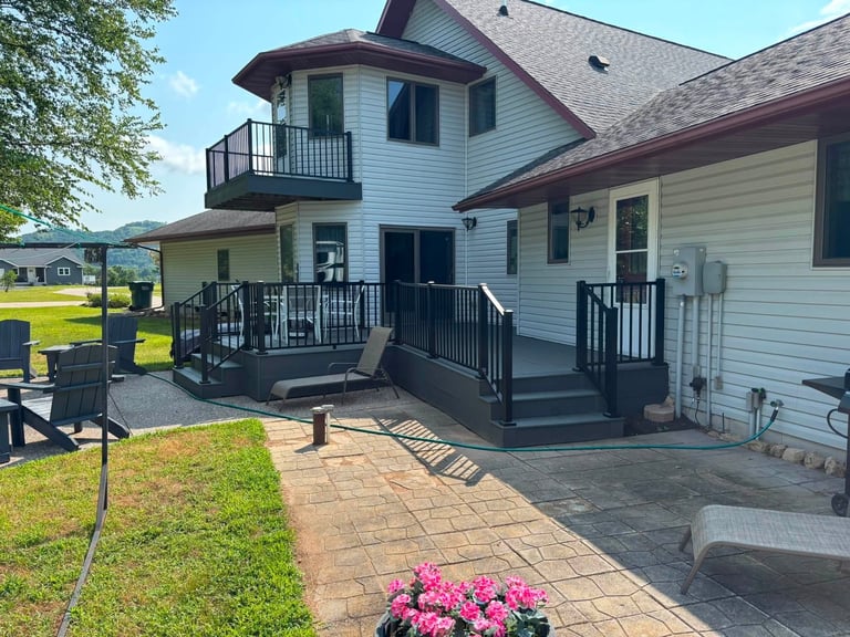 Two-story white house with black deck and railings, brick patio, pink flowering plants, and green lawn on sunny day