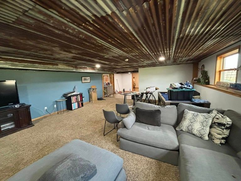 Spacious basement recreation room with gray sectional sofa, corrugated metal ceiling, light blue accent wall, and carpeted floor
