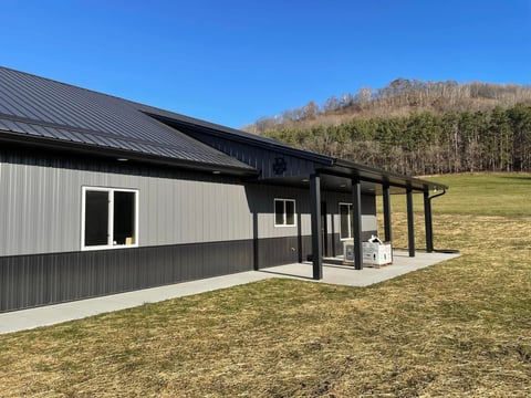 Modern metal-sided house with black roof and covered porch in rural hillside setting with clear blue sky