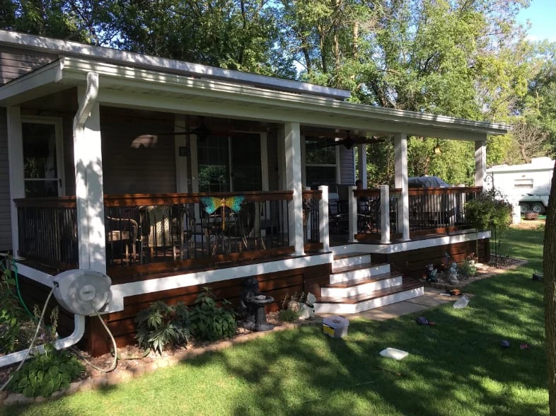Charming single-wide mobile home with covered porch, colorful chairs, brick steps, and mature trees in yard