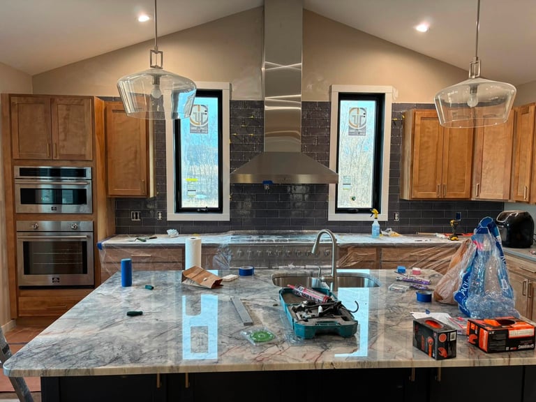 Modern kitchen undergoing renovation with tools and materials scattered across marble countertop, displaying wooden cabinets and stainless steel appliances.
