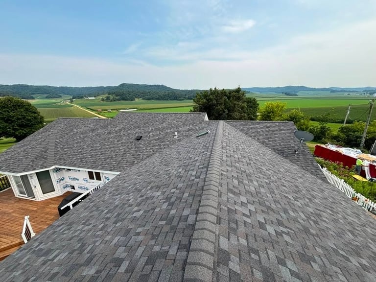 Aerial view of a residential roof with gray asphalt shingles overlooking green farmland and countryside landscape