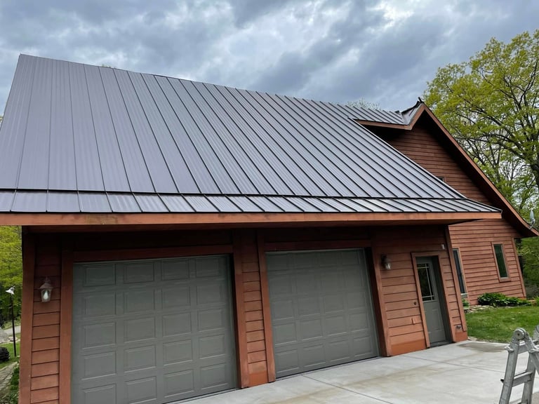 Modern residential garage with reddish-brown siding, metal roof, two gray garage doors, and green trees