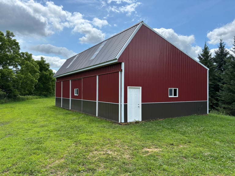 Red metal barn with gray base and white trim doors, set in grassy field with trees and cloudy sky