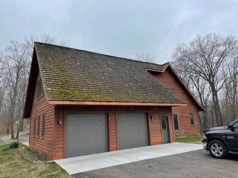 Red brick garage building with two bay doors, peaked roof with moss, surrounded by bare trees and parked vehicle