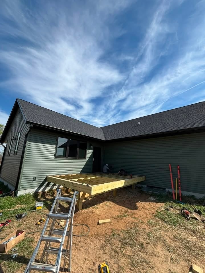 Under-construction deck attached to a dark-colored house with wooden framing, aluminum ladder, and tools visible in yard