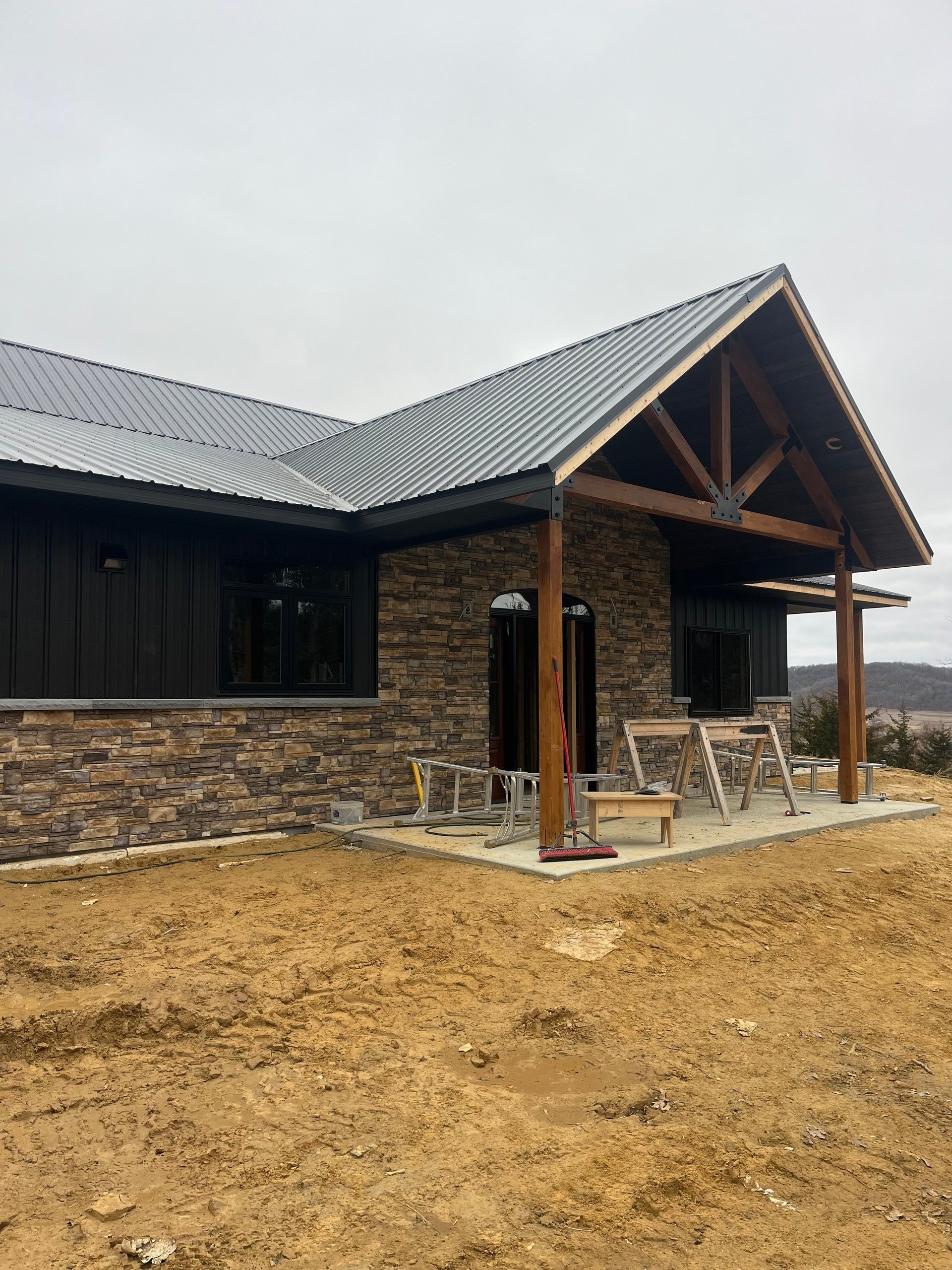 Modern house with stone facade, metal roof, wooden covered porch, and metal chairs on bare earth yard