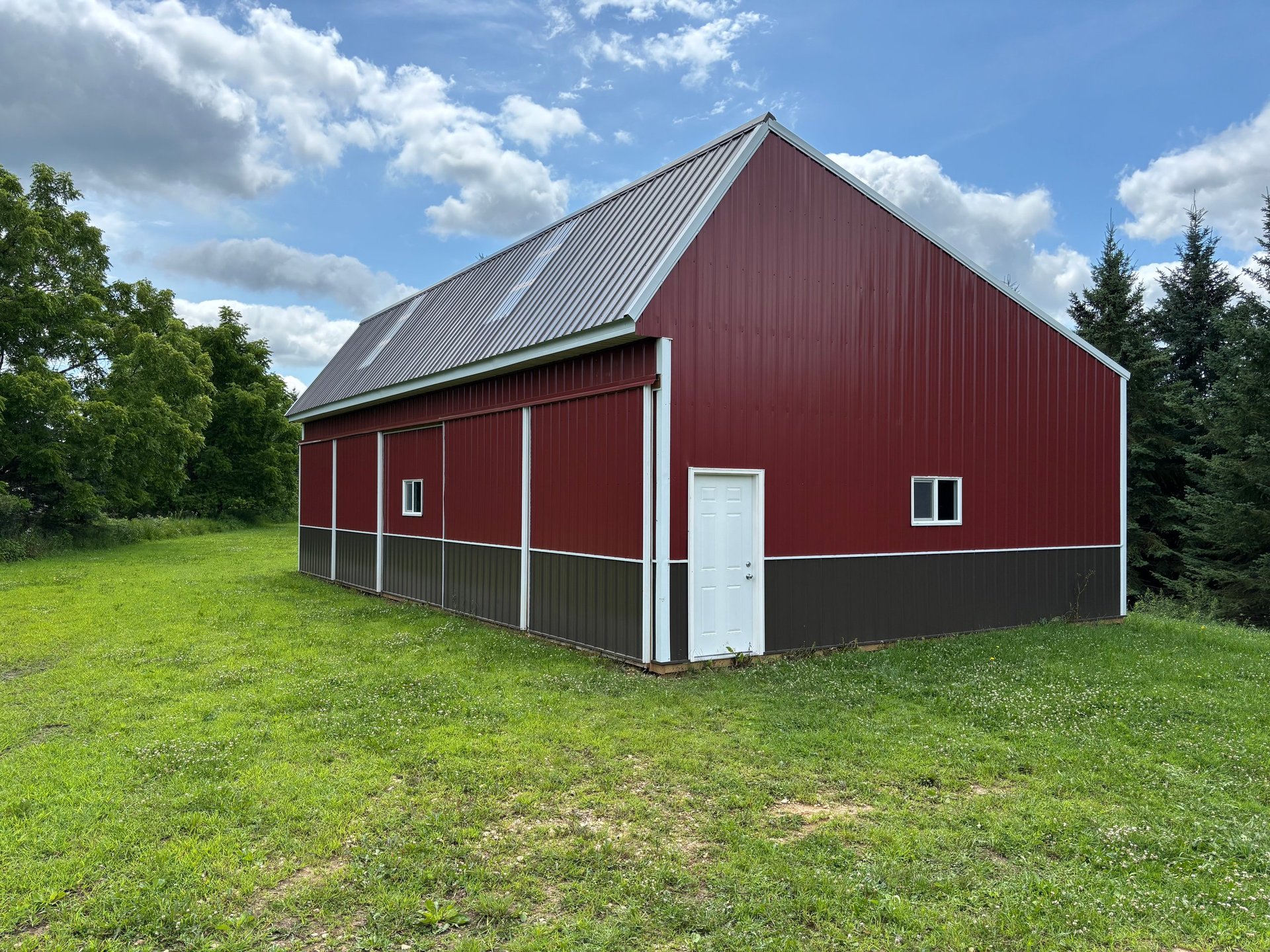 Red metal barn with metal roof on grassy field surrounded by trees under blue sky with white clouds