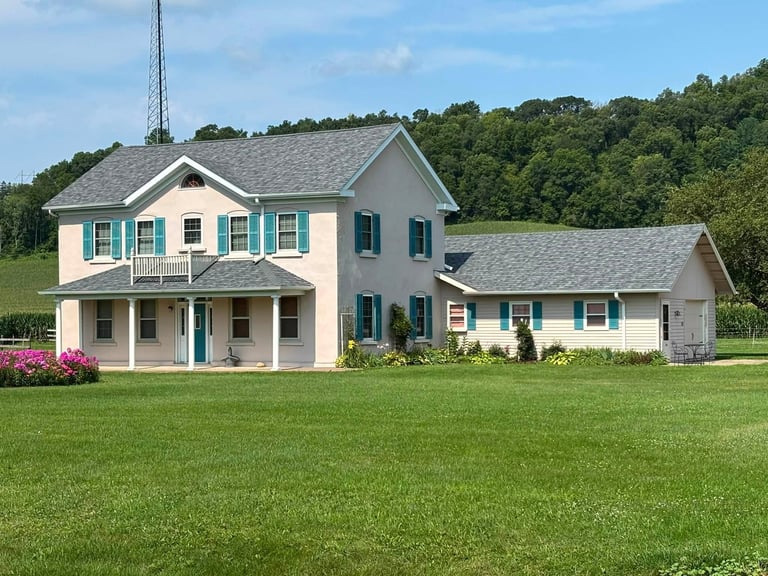 Large two-story farmhouse with cream and blue exterior, front porch, and attached garage on green lawn with wooded background