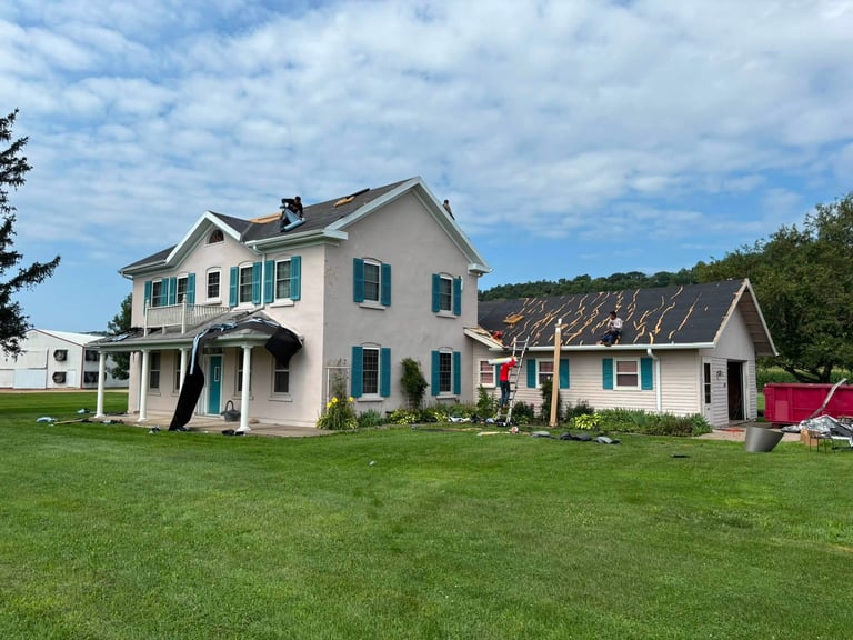 White farmhouse with blue shutters and detached garage on green lawn with wooded hills in background