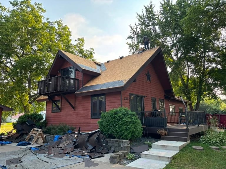 Red wooden house with peaked roof, wraparound porch, and manicured landscaping surrounded by mature trees