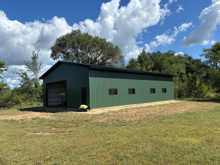 Green metal barn with dark roof on grassy property under blue sky with white clouds