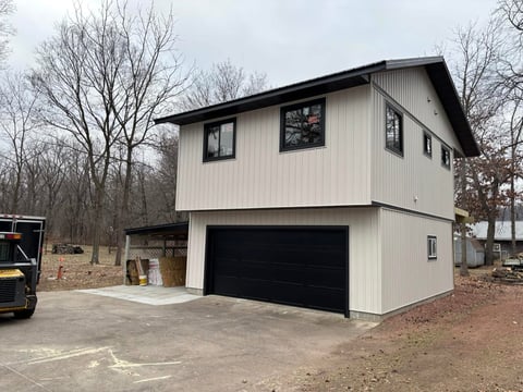 White two-story modern garage building with black roof and large black garage door on bare dirt lot surrounded by bare trees