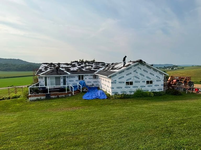 Damaged house with roof destroyed, blue tarp covering debris, sitting in open green field with hills in background