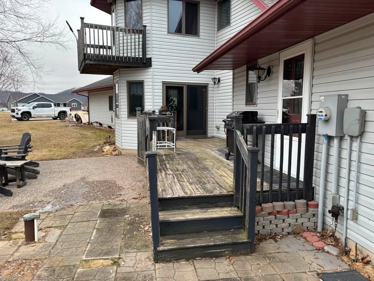 Exterior of two-story white house with wooden deck, black railing, and brick patio area, parked white vehicle visible in driveway.