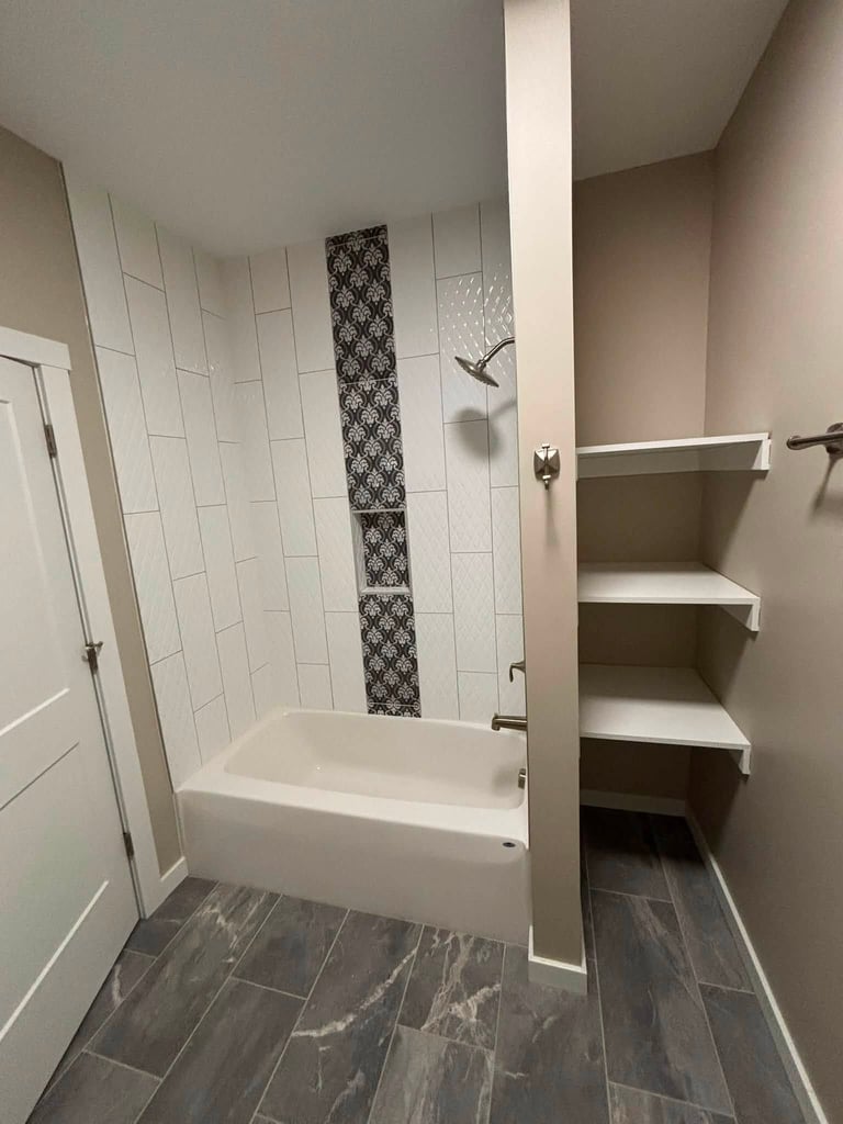 Modern bathroom with white subway tile shower surround, decorative black accent strip, white soaking tub, built-in shelves, and dark wood-look flooring